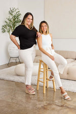 Two women wearing white jeans, posing together in a modern living room setting.