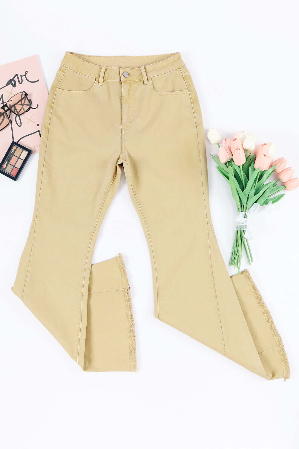 Beige pocketed raw hem flare jeans displayed with a bouquet of pink tulips and a makeup palette on a white background