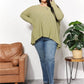 Woman wearing oversized super soft rib layering top with sharkbite hem and round neck, styled with jeans and sandals, standing by armchair and plant.