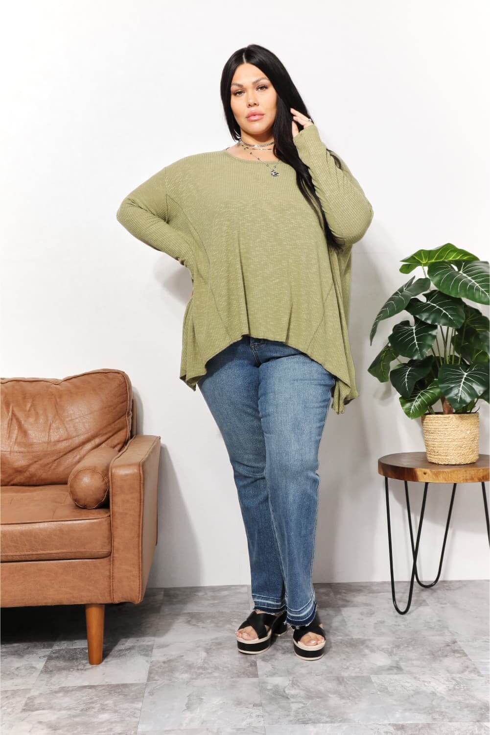 Woman wearing oversized super soft rib layering top with sharkbite hem and round neck, styled with jeans and sandals, standing by armchair and plant.