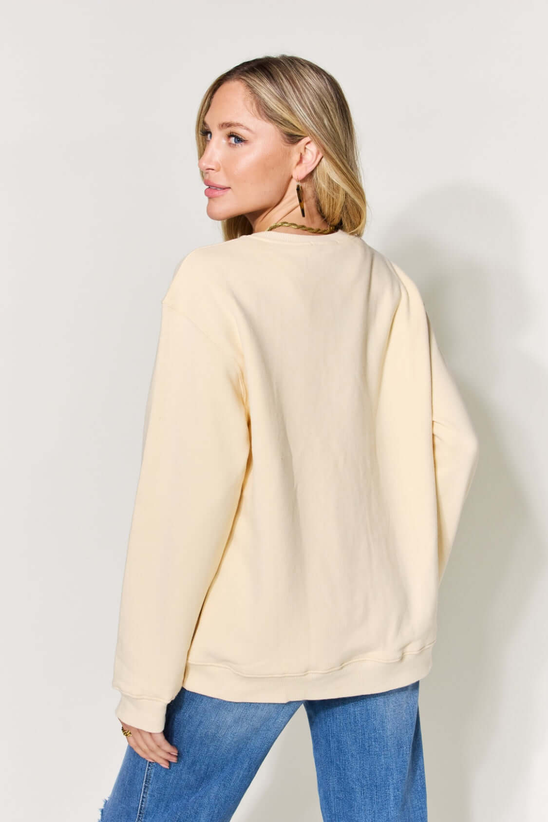 Woman wearing Letter Graphic Long Sleeve Sweatshirt in beige, showing back view. Styled with blue denim jeans.