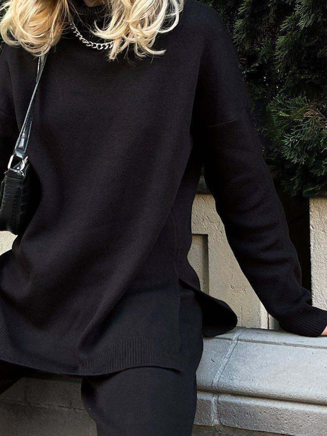 Woman wearing black side slit turtleneck sweater with dropped shoulders, styled with a chain necklace and small black bag.