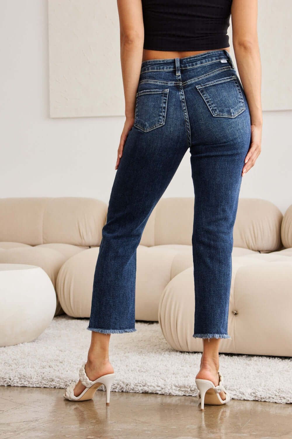 Woman wearing RFM Jeans Crop Dylan Full Size Tummy Control Distressed High Waist Raw Hem Jeans, stylish backside view in modern living room