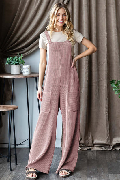 Woman wearing Ribbed Front Pocket Sleeveless Jumpsuit, standing indoors with plants and a brown curtain backdrop