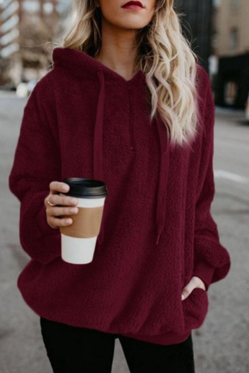 Stylish woman wearing a burgundy teddy hoodie with drawstrings, holding a coffee cup on a city street.