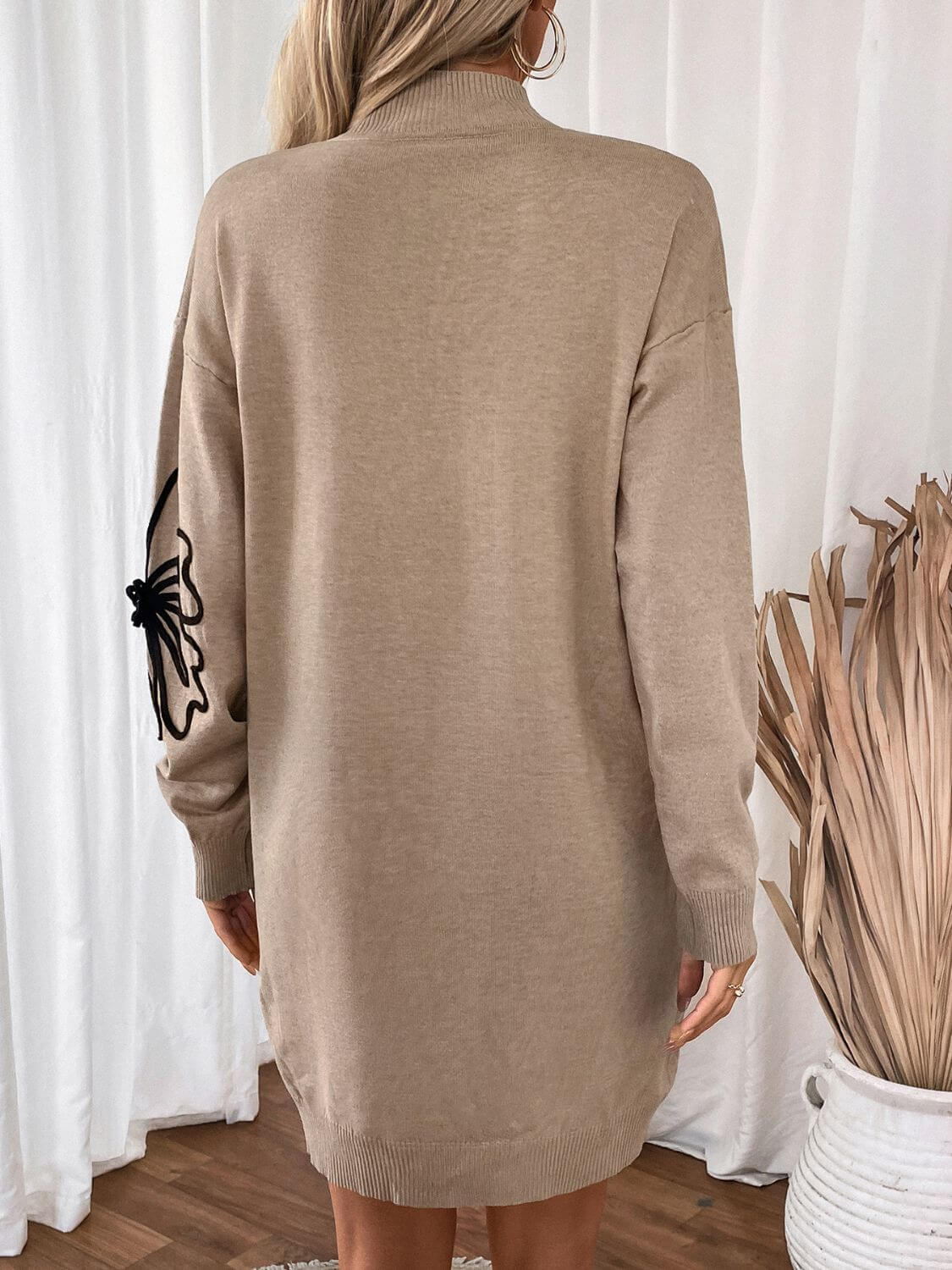 Back view of Perfee Flower Mock Neck Long Sleeve Sweater Dress in tan with flower detail on sleeve, wooden floor and white curtains in the background