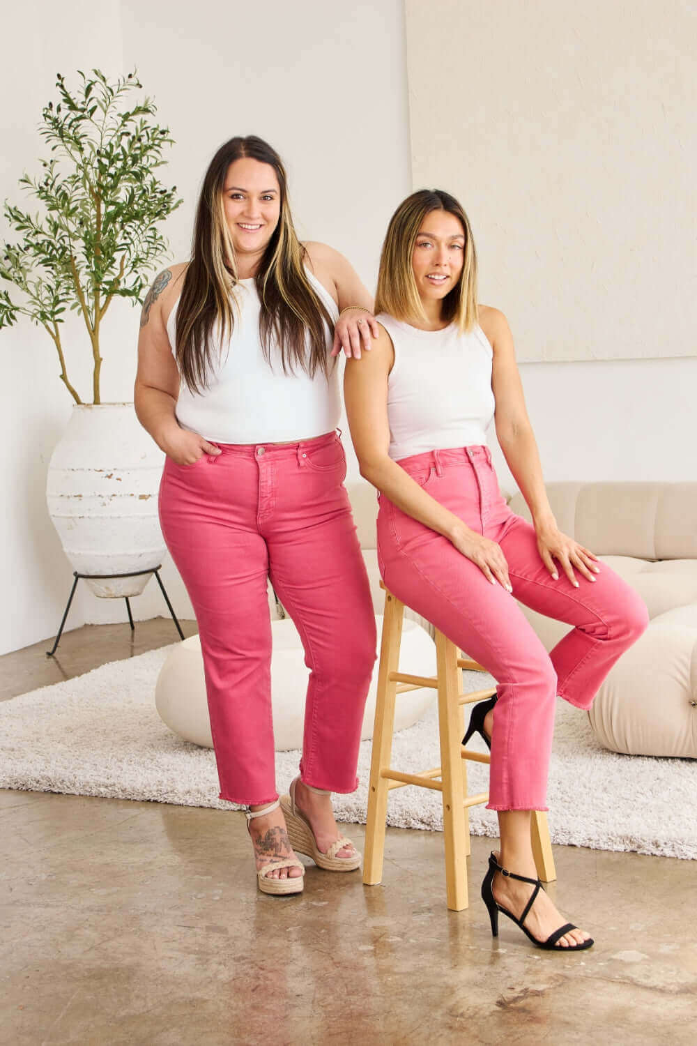 Two women wearing pink high-waisted jeans and white tops, showcasing RFM Jeans with a stylish raw hem design in a modern and comfortable setting.