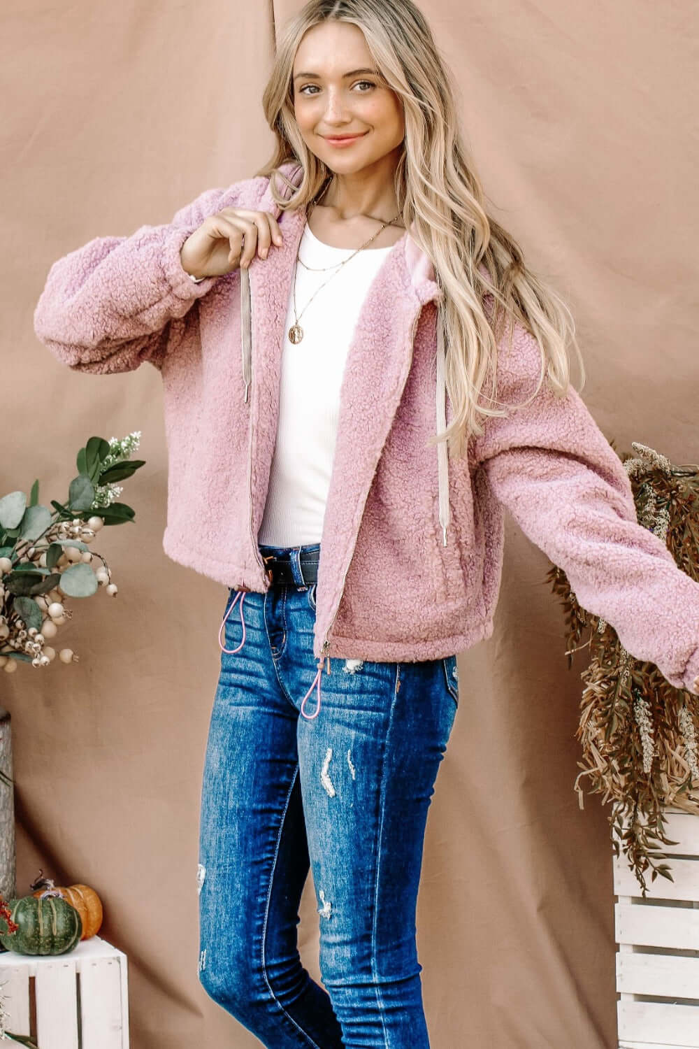 Woman wearing And The Why Sherpa Drawstring Cropped Hooded Jacket in pink, standing outdoors, pairing it with blue jeans.