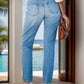 Woman wearing distressed buttoned jeans with pockets, standing by a poolside patio, holding a brown handbag.