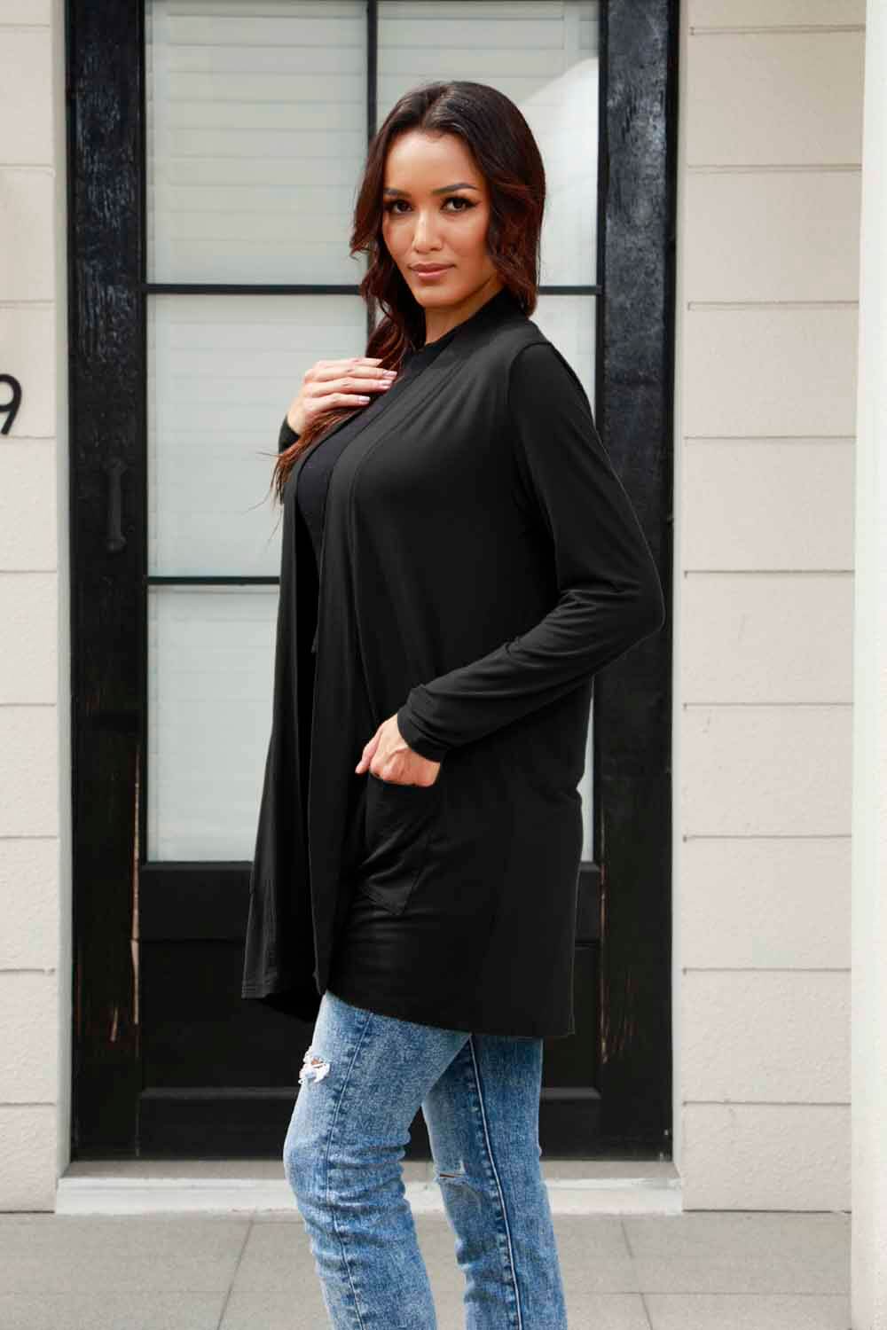 Woman wearing black open front long sleeve cardigan with pockets over blue jeans standing in front of a door