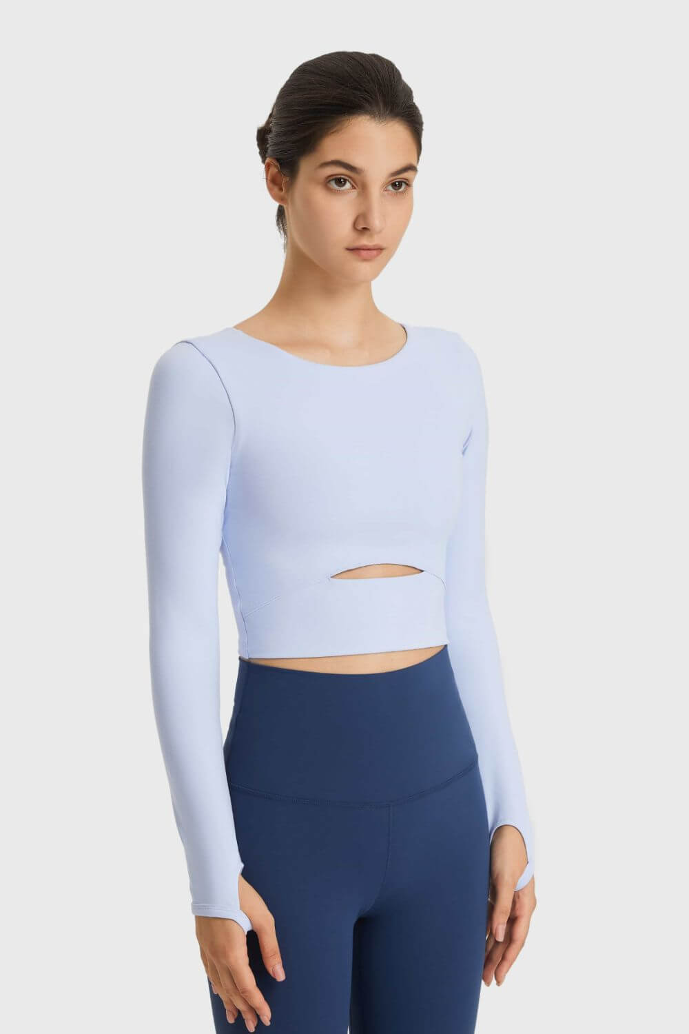 Stylish model wearing Millennia Cutout Long Sleeve Cropped Sports Top in light blue, paired with navy leggings in a studio.