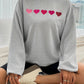 Woman wearing Devine Heart Dropped Shoulder Long Sleeve Sweatshirt with heart design while sitting on a bed.