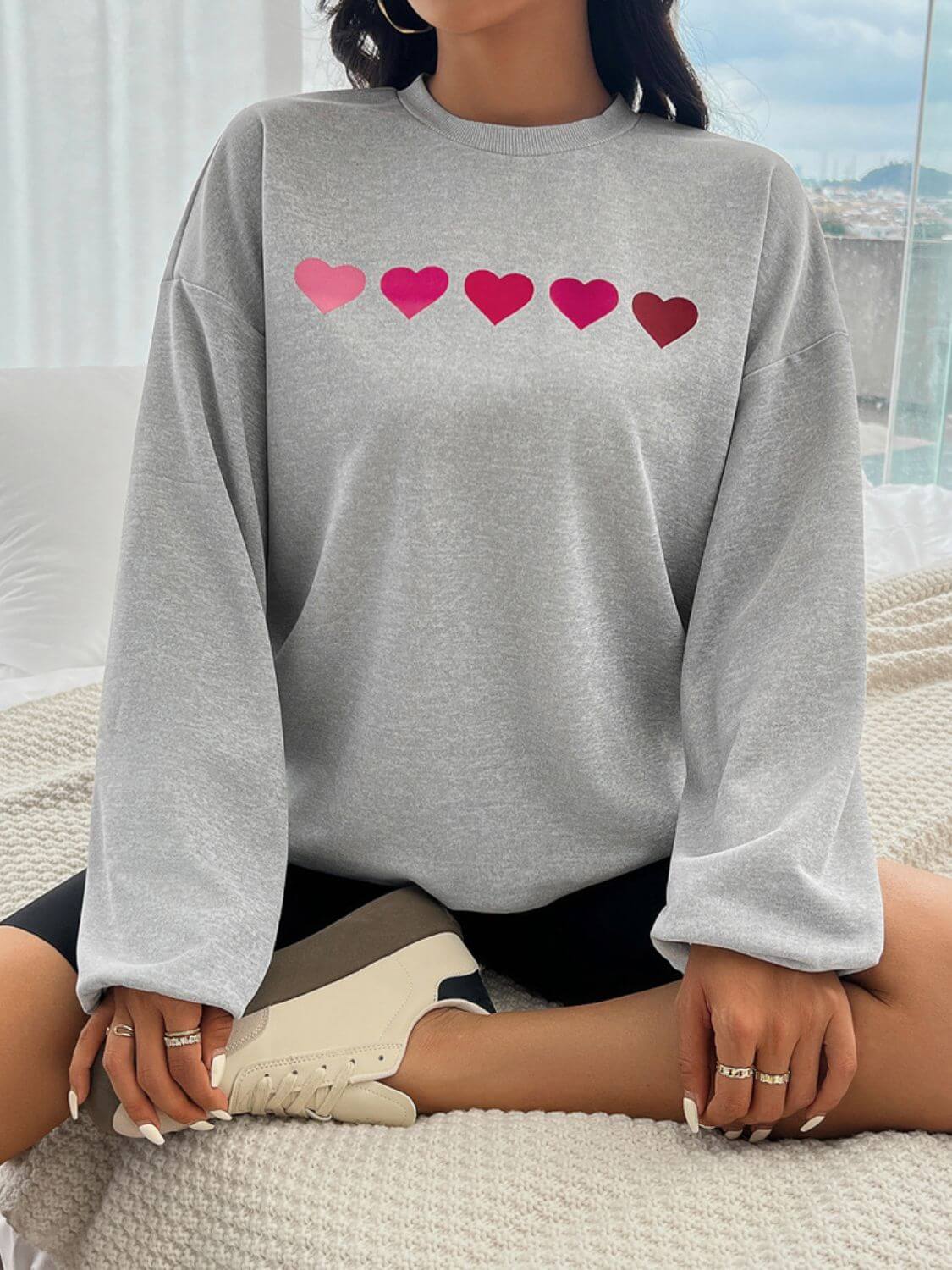 Woman wearing Devine Heart Dropped Shoulder Long Sleeve Sweatshirt with heart design while sitting on a bed.