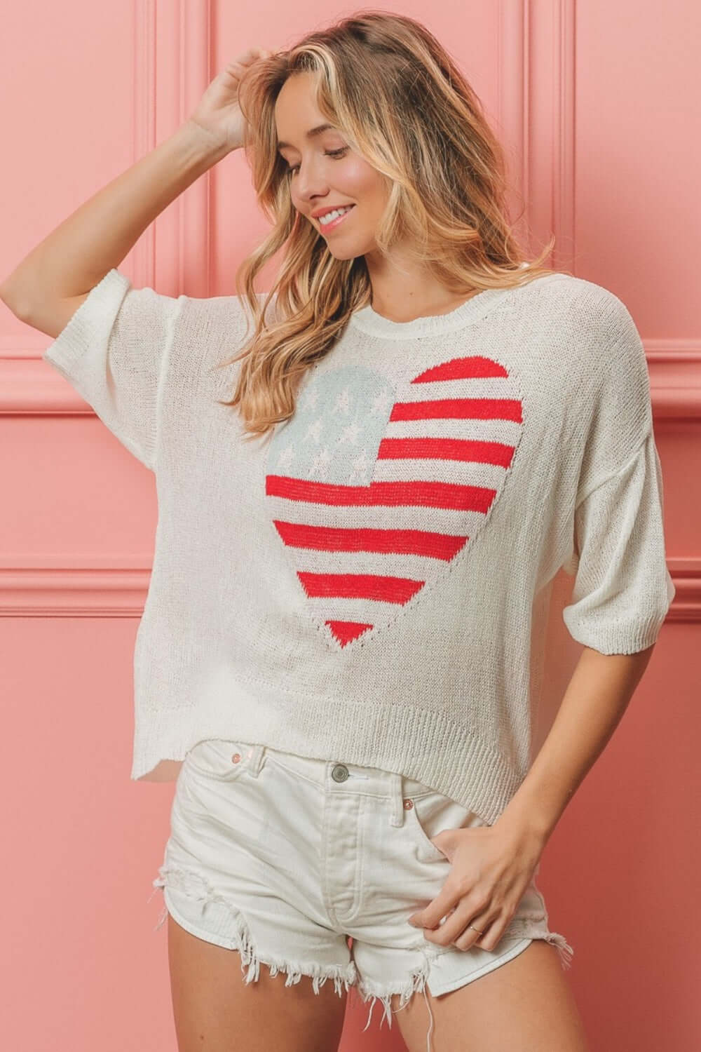 Woman wearing a cozy knit striped heart contrast top with red and white stripes, paired with white shorts, against a pink background.