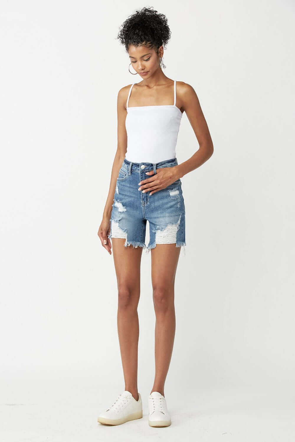 Model wearing high-waisted distressed denim shorts by Risen Jeans with a white tank top and sneakers for a casual summer look