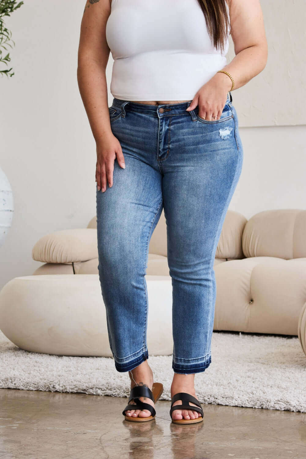 Woman wearing Judy Blue release hem cropped bootcut jeans standing in a stylish living room with white furniture.