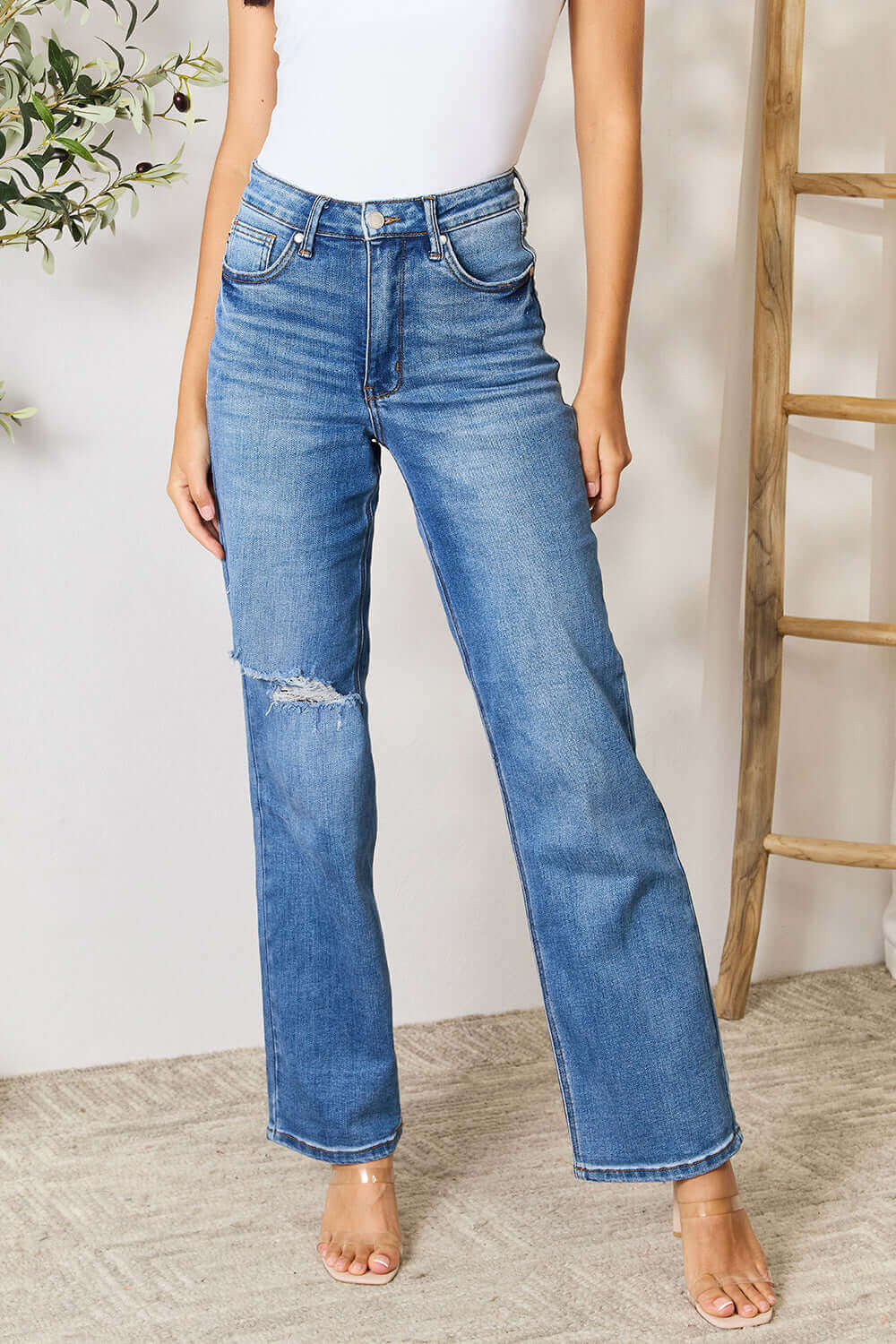 Woman wearing High Waist Distressed Judy Blue Jeans Full Size with a white top standing in a stylish indoor setting