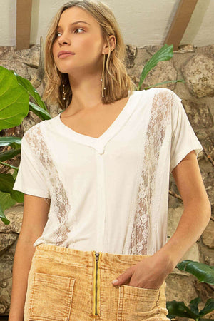 Woman wearing a white short sleeve V-neck T-shirt with lace outseam detail and relaxed fit, paired with mustard yellow pants.