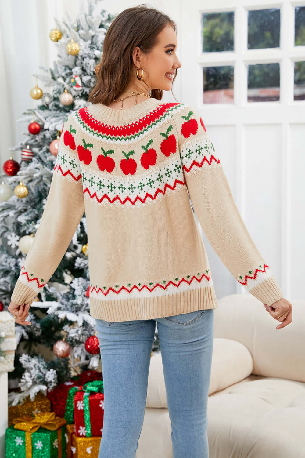 Woman wearing an angel wings sweater with apple design, round neck, and long sleeves, standing by a decorated Christmas tree.