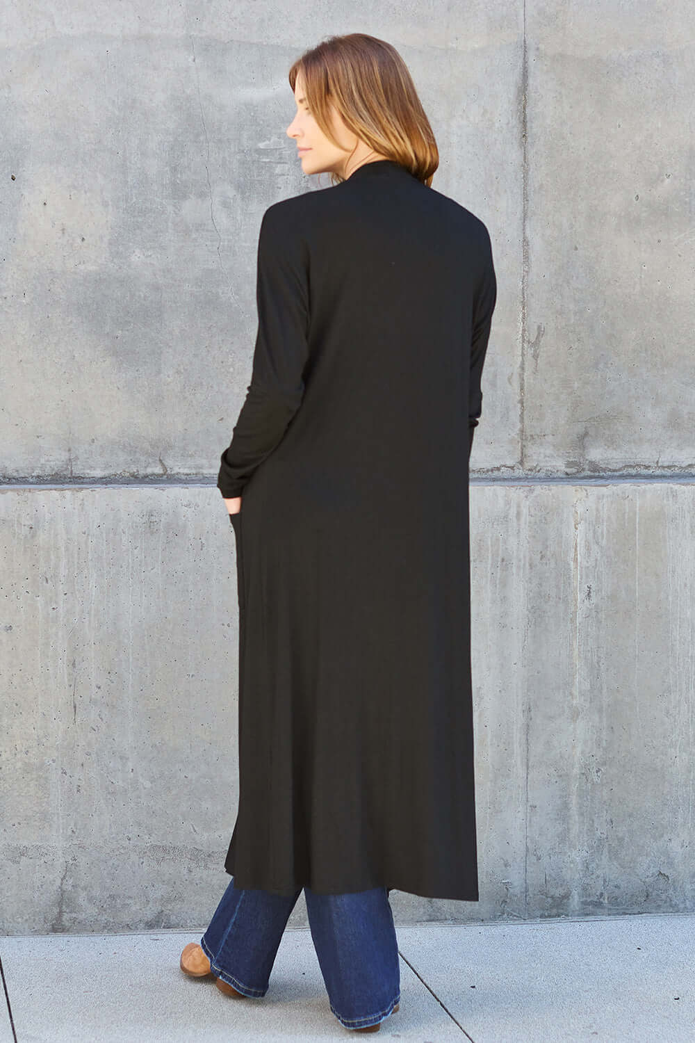 Woman wearing a black open front long sleeve cover up with pockets, jeans, and brown shoes, standing against a gray concrete wall.