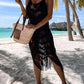 Woman in a black fringe cover-up poses by the beach with a straw bag, showcasing a chic poolside style.