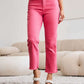 Woman wearing Crop Dylan Full Size Tummy Control High Waist Raw Hem RFM Jeans in pink standing in a stylish room.