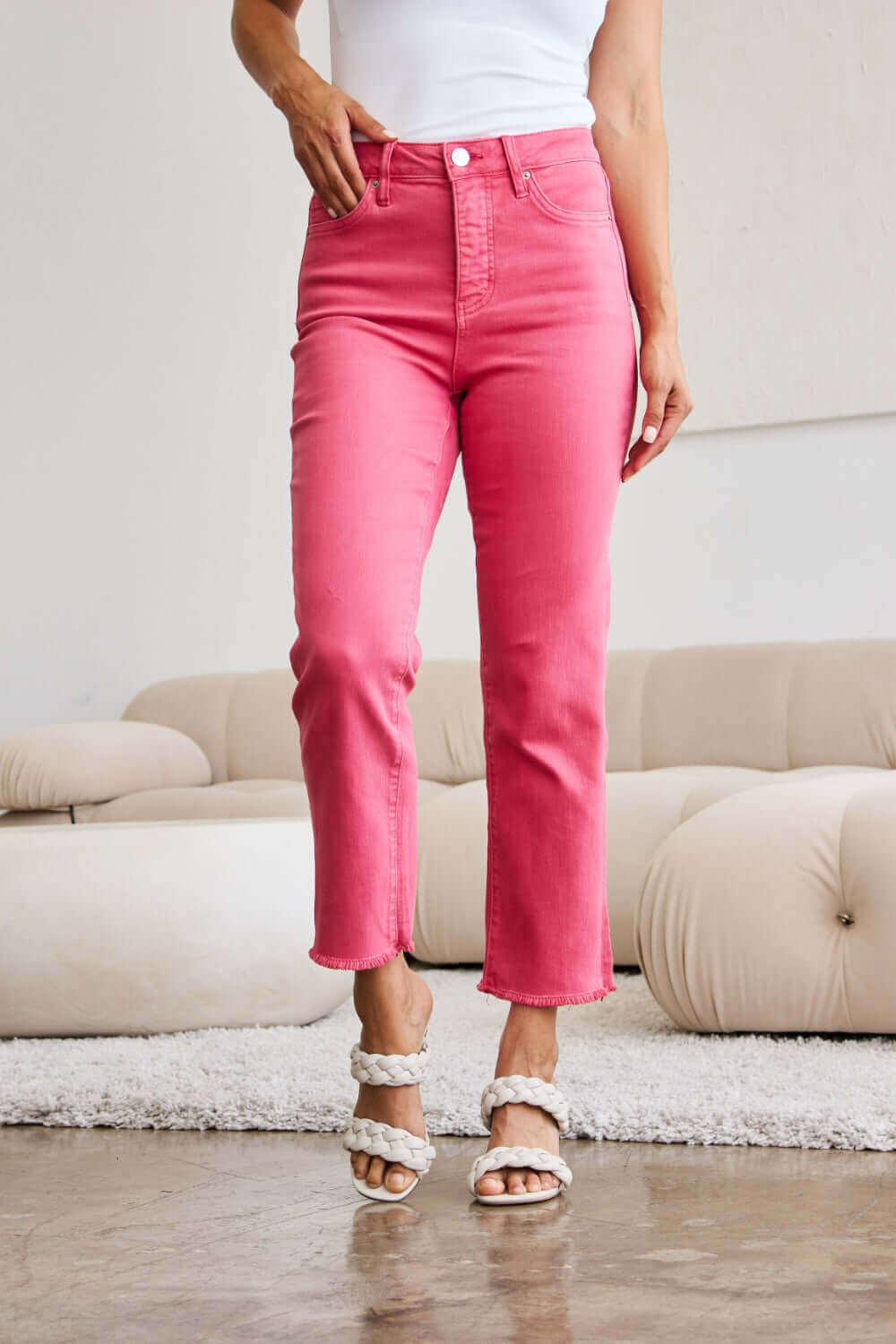 Woman wearing Crop Dylan Full Size Tummy Control High Waist Raw Hem RFM Jeans in pink standing in a stylish room.