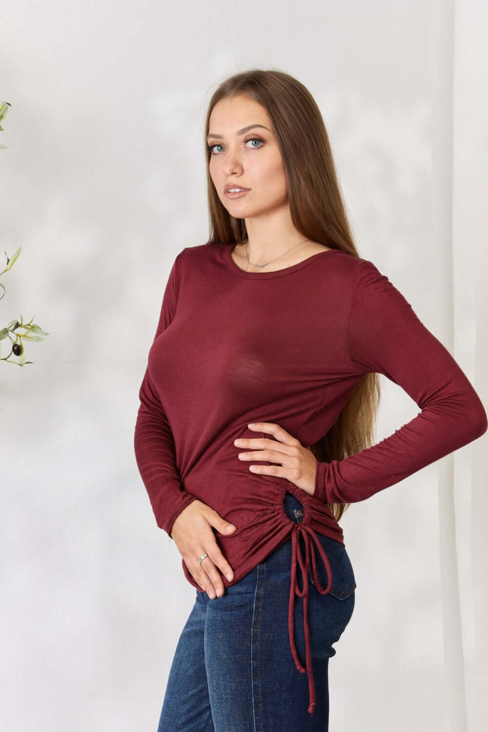 Woman wearing a burgundy drawstring round neck long sleeve top, paired with blue jeans, posing confidently against a neutral background.