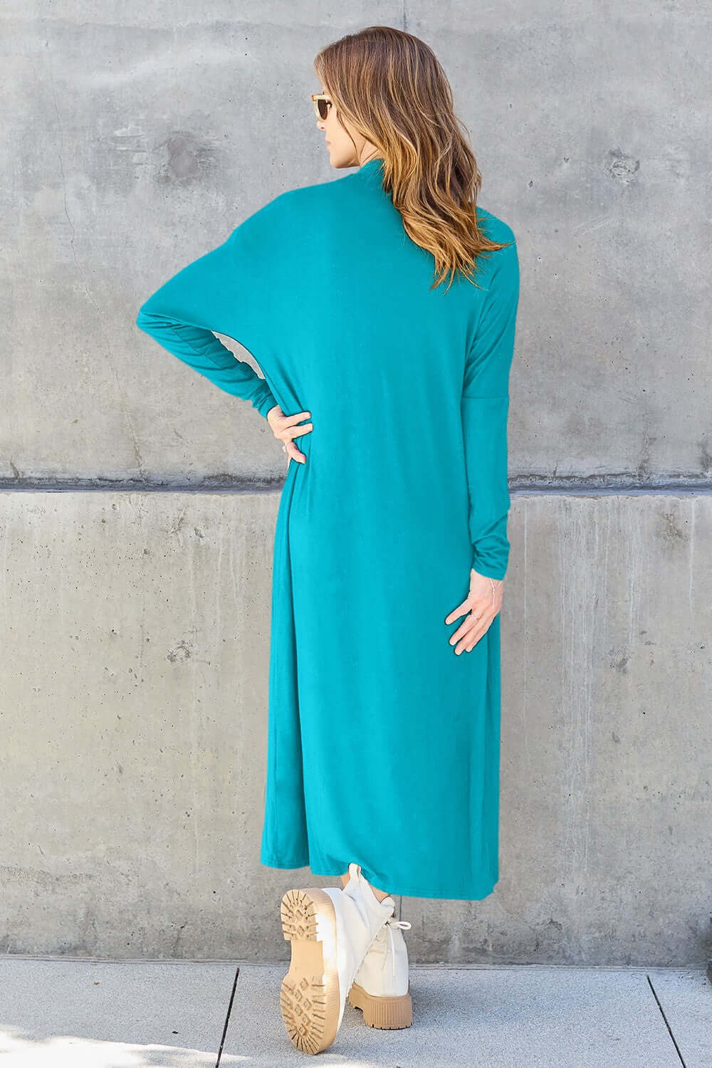 Woman wearing turquoise open front long sleeve cover up with slightly stretchy material, seen from the back against a concrete wall