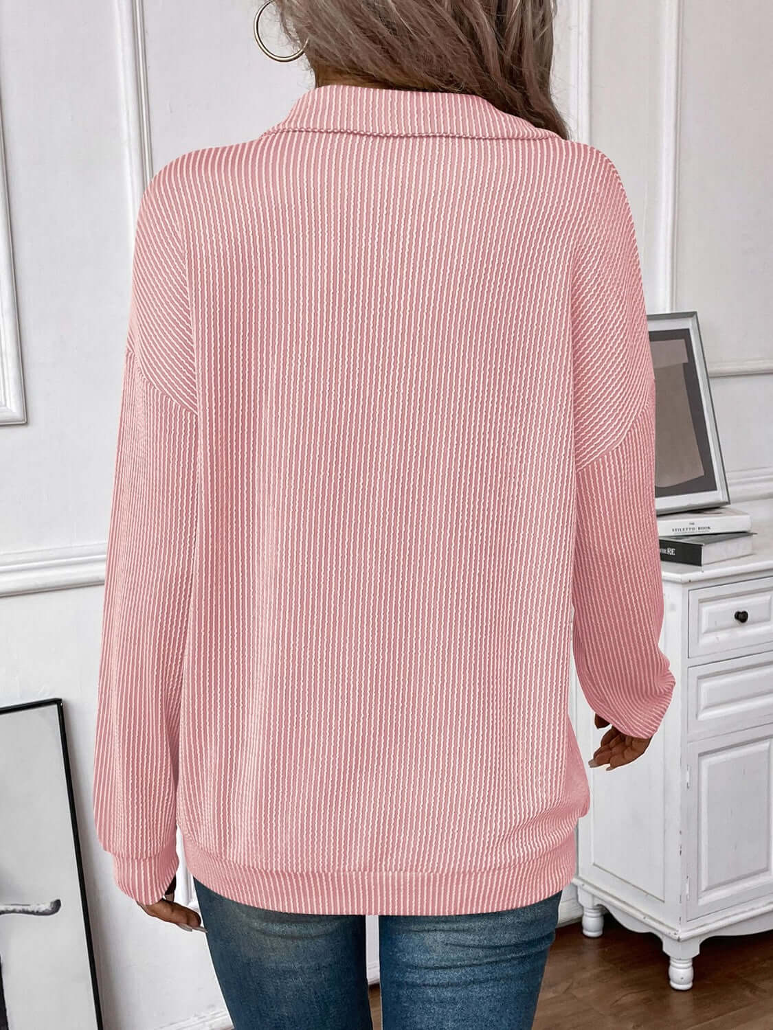 Back view of a woman wearing a striped half zip long sleeve pink T-shirt, standing in a white room with desk and computer in the background.