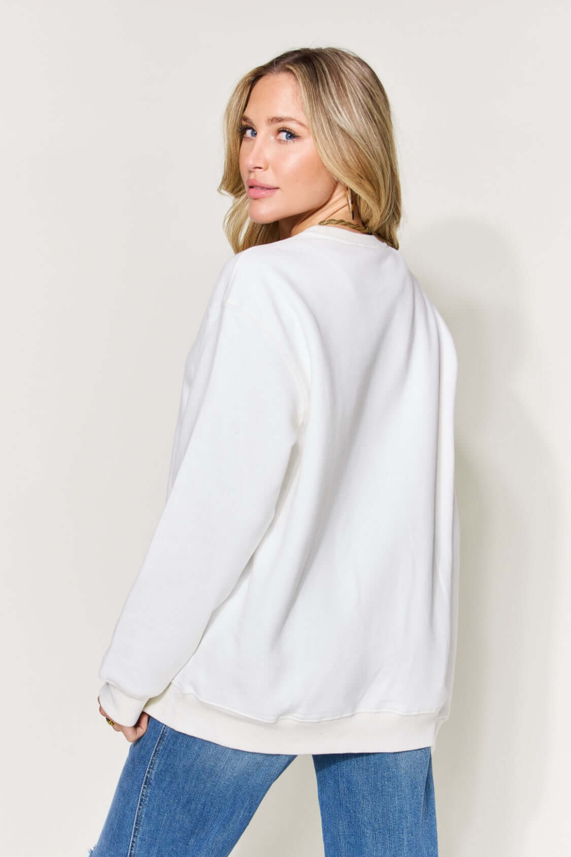 Woman wearing a white long sleeve sweatshirt with letter graphic on back, paired with blue jeans, showcasing the back view.