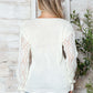 Woman wearing Bella Road openwork V-neck long sleeve sweater in white, showcasing stylish design against rustic backdrop.