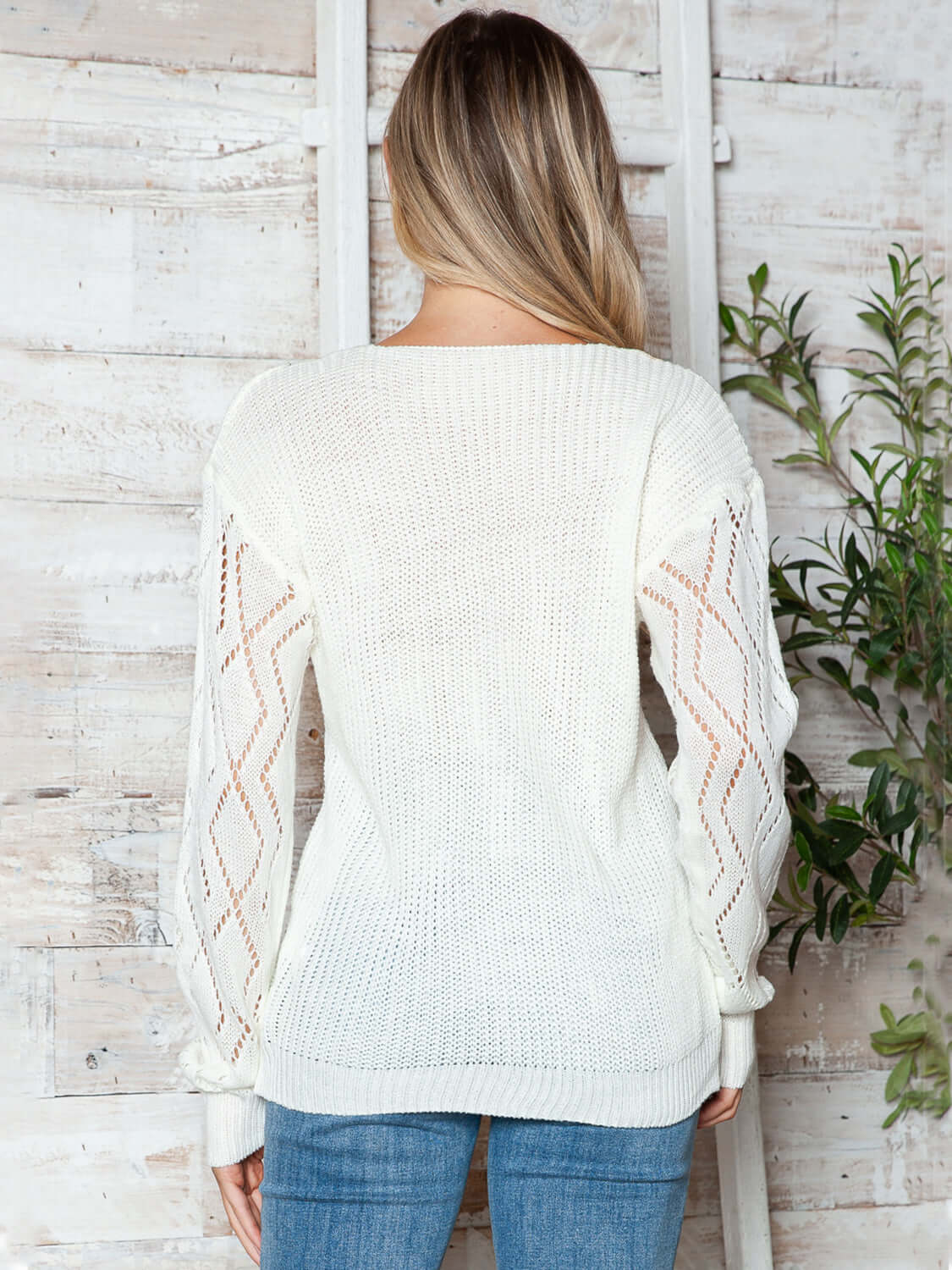 Woman wearing Bella Road openwork V-neck long sleeve sweater in white, showcasing stylish design against rustic backdrop.