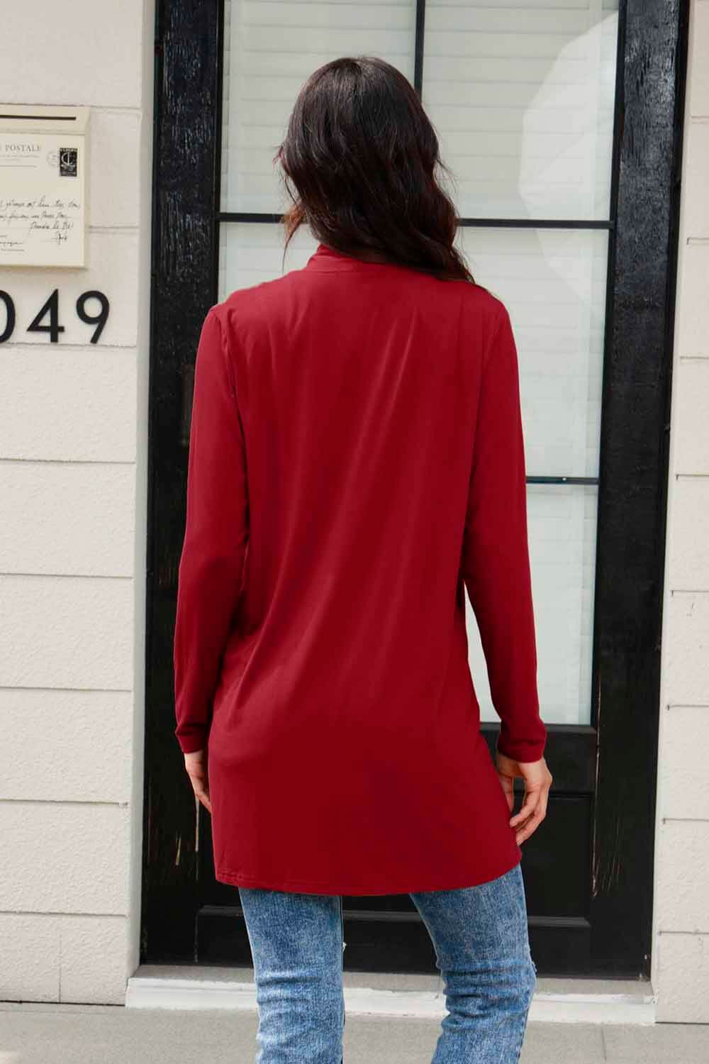 Woman wearing red open front long sleeve cardigan with pockets, standing in front of a black door, showcasing back design.