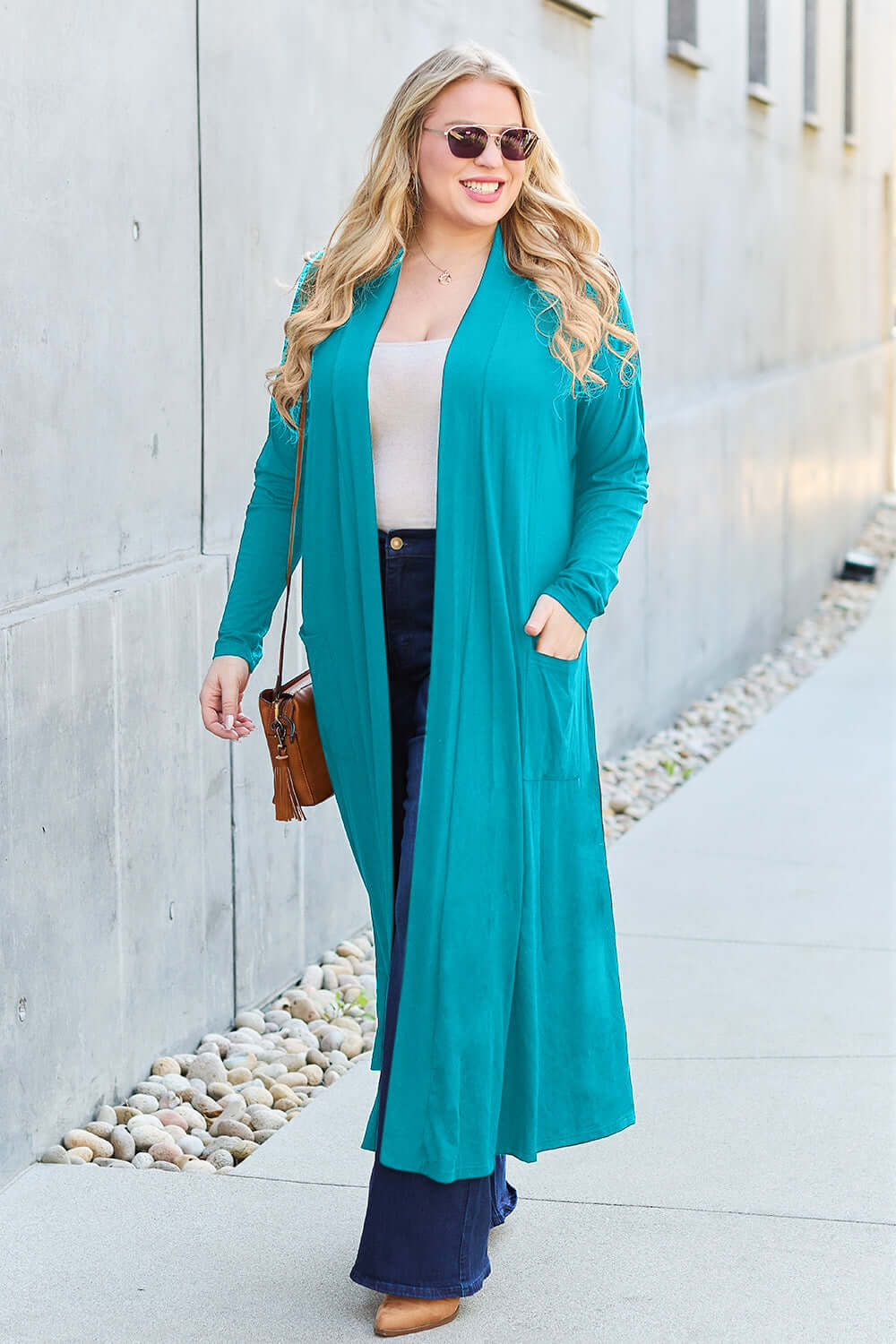 Woman wearing teal open front long sleeve cover up with pockets, paired with white top and blue jeans, walking outdoors.