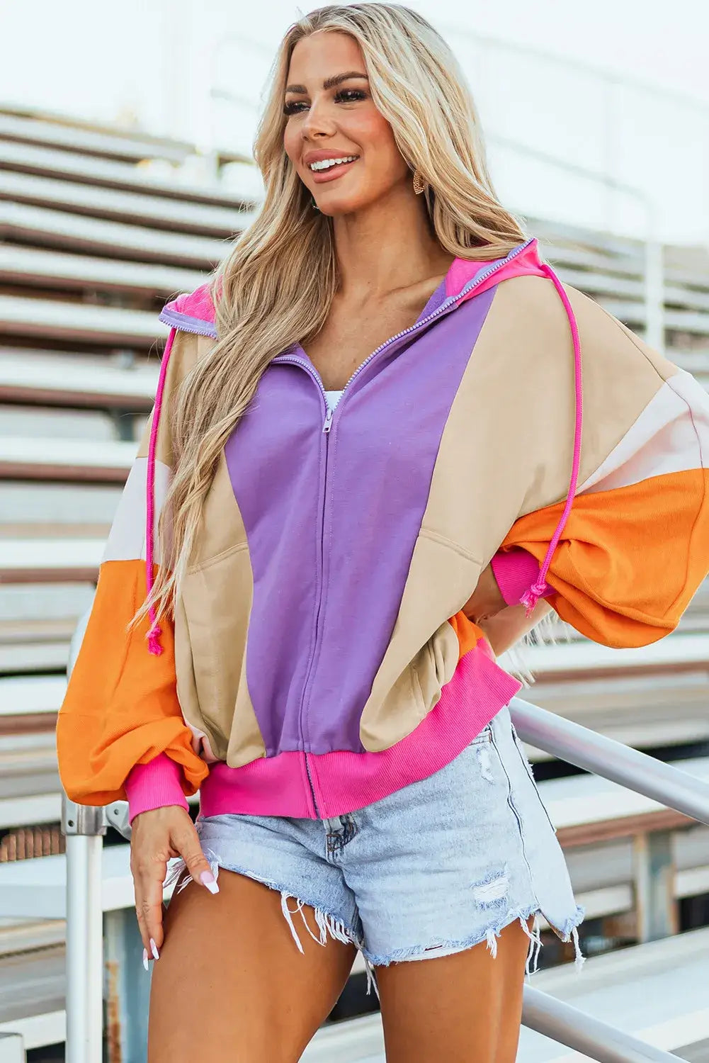 Woman wearing drawstring color block zip up long sleeve hoodie in purple, beige, orange, and pink, paired with denim shorts.