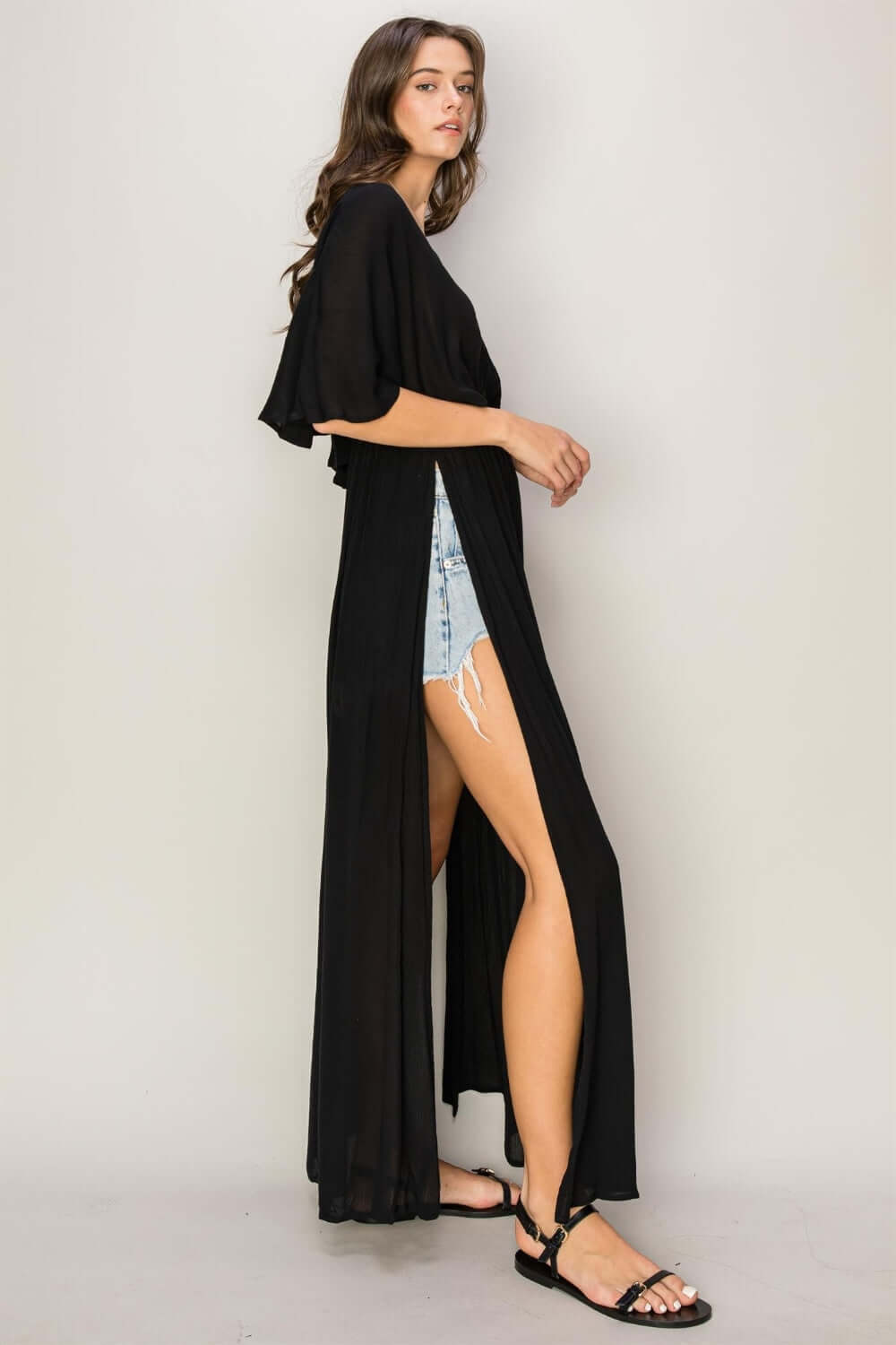 Woman wearing tie back maxi split cover-up dress with a plunging neck, batwing sleeves, and side slits showing denim shorts.