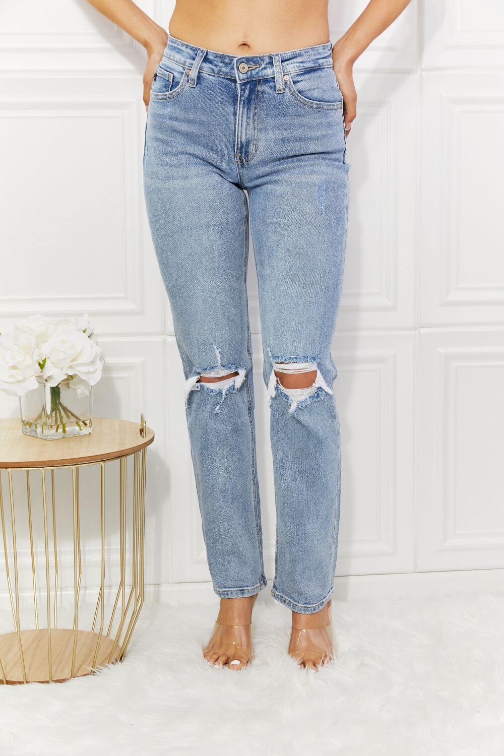 High rise distressed slim straight jeans with ripped knees in light wash, featuring pockets and slight stretch, worn by a model, next to a table with flowers.