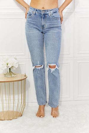 High rise distressed slim straight jeans with ripped knees in light wash, featuring pockets and slight stretch, worn by a model, next to a table with flowers.