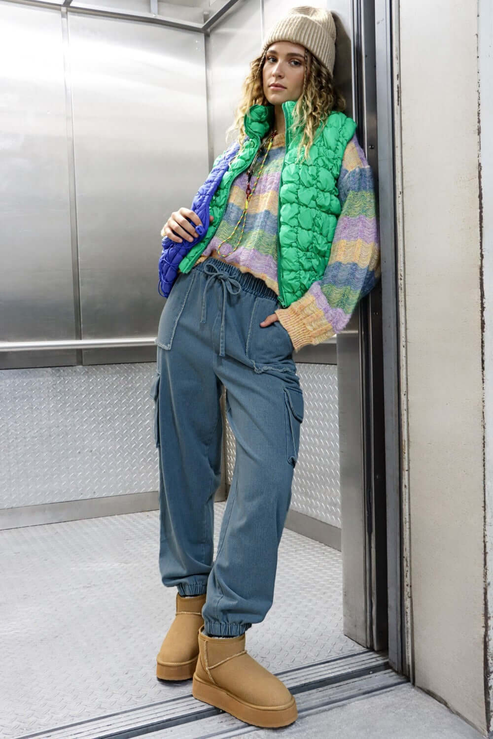 Woman wearing washed drawstring jogger cargo jeans with pockets, paired with a colorful sweater and puffer vest in an elevator.