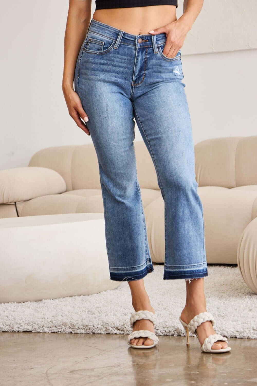 Woman wearing Release Hem Cropped Bootcut Judy Blue Jeans styled with white heels in a modern living room.