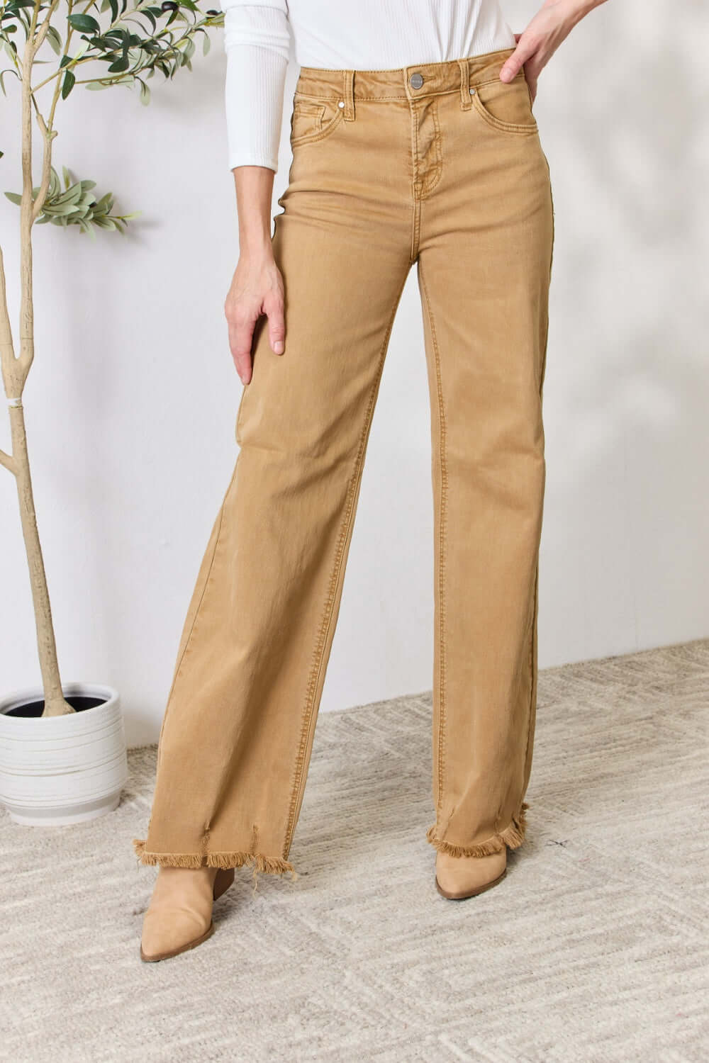 Woman wearing high-waisted fringe hem wide leg Risen Jeans in tan, standing next to a potted plant in a stylish, neutral room.