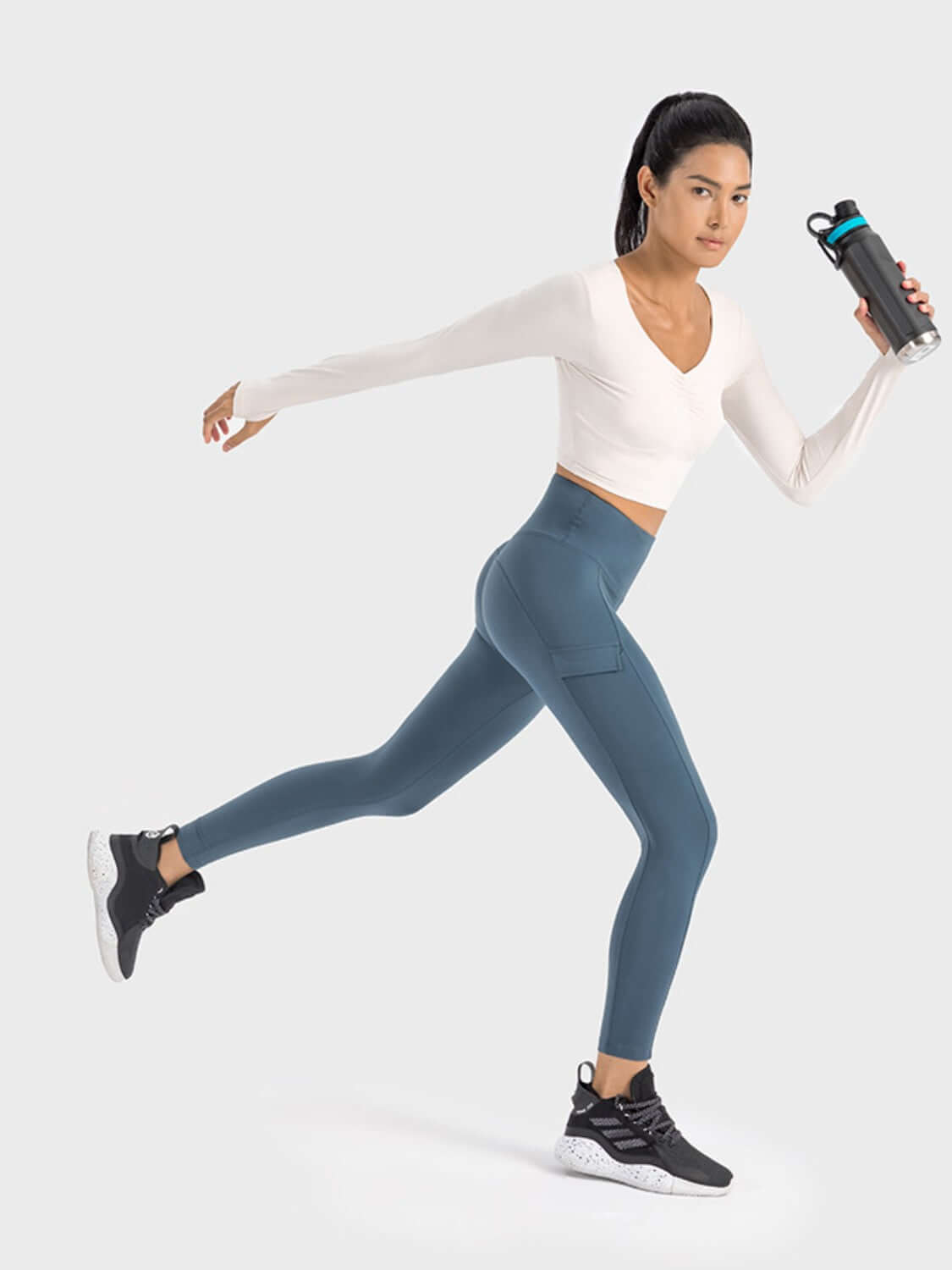 Model wearing a Millennia Ruched Cropped Long Sleeve Sports Top and leggings, holding a water bottle, showcasing active lifestyle.