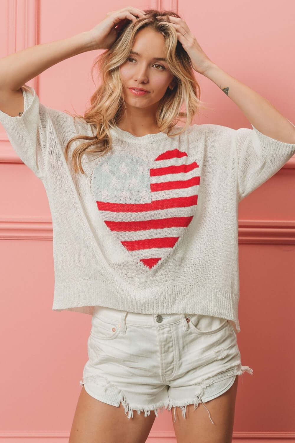 Woman wearing striped heart contrast knit top with distressed white shorts in front of a pink wall