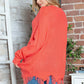 Vibrant orange sweater with frayed hem on woman against rustic brick wall backdrop.