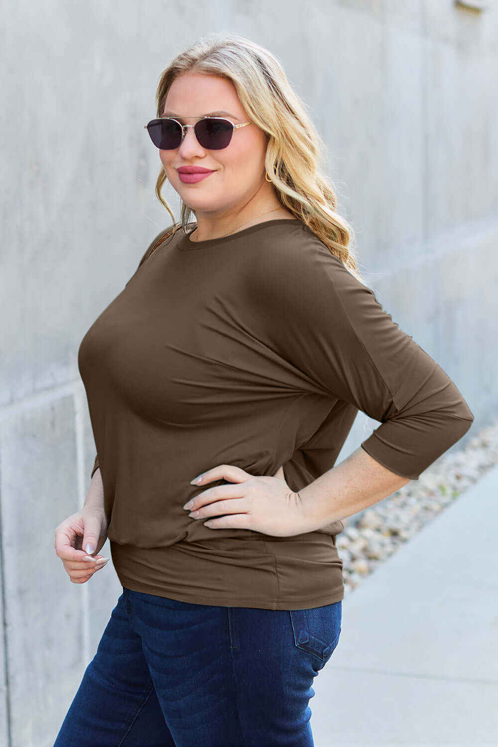 Woman wearing a round neck batwing sleeve top in brown, paired with blue jeans, posing with one hand on hip outdoors.