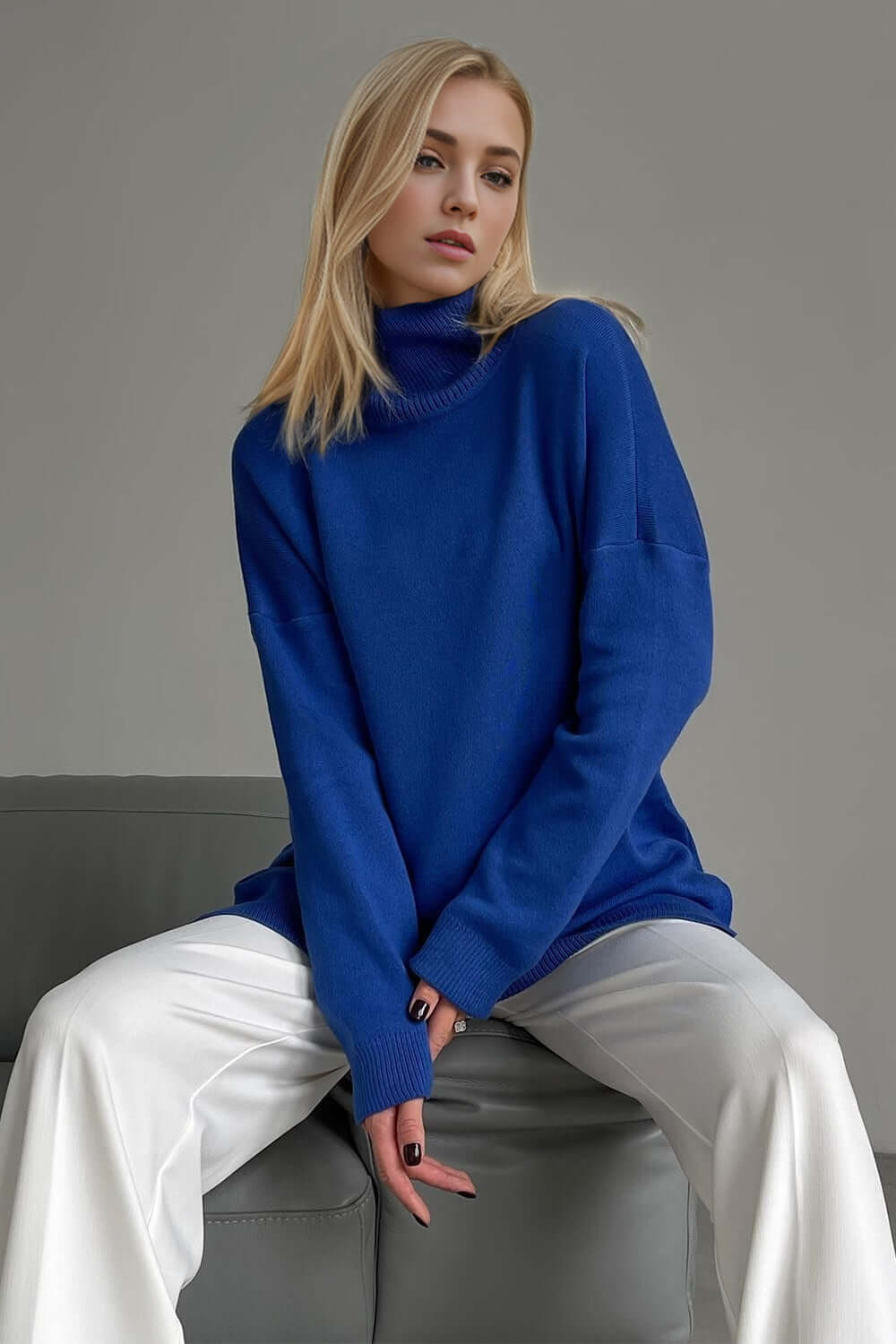 Woman in a blue turtleneck sweater with side slits and dropped shoulders, seated stylishly on a gray couch.