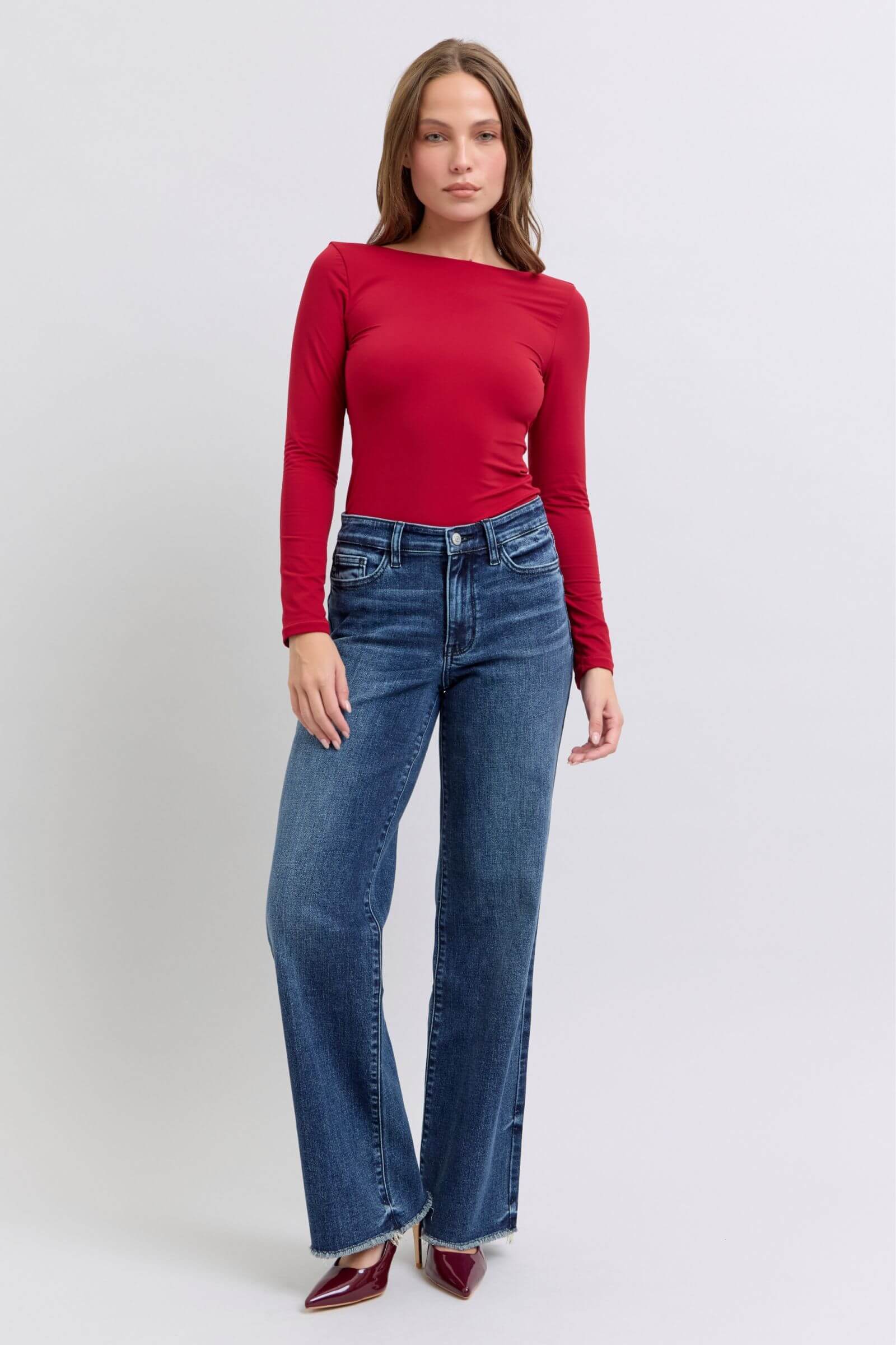 Model wearing Judy Blue Full Size Raw Hem Mid Rise Jeans with a fitted red long-sleeve top, showcasing a stylish casual look.