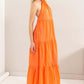 Model wearing an orange halter neck cover up maxi dress with a tiered silhouette and open back design.