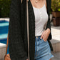 Woman wearing a black openwork open front dropped shoulder cardigan, white tank top, denim shorts, and a brown handbag by the pool.